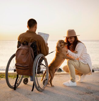 A young man in a wheelchair and a woman in hat playing with a dog