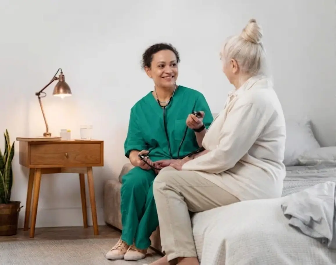 A smiling home care nurse in green scrubs checking the blood pressure of an elderly woman sitting on a bed.