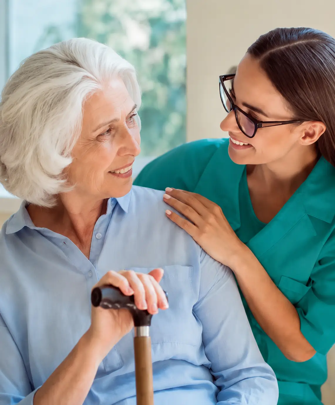 A smiling female nurse in teal scrubs comforting an elderly senior woman holding a walking cane, representing home health care and assisted living services.