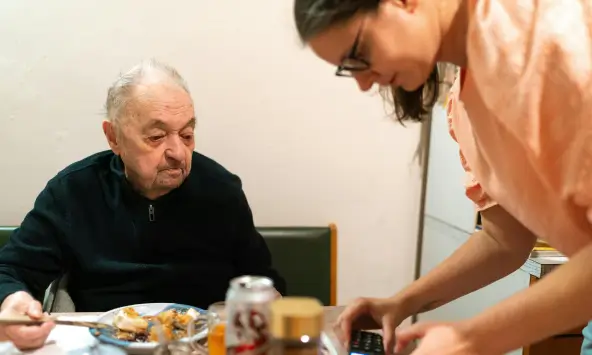 A caregiver providing senior home care in Toronto by assisting an elderly man at the dining table with a handheld device.