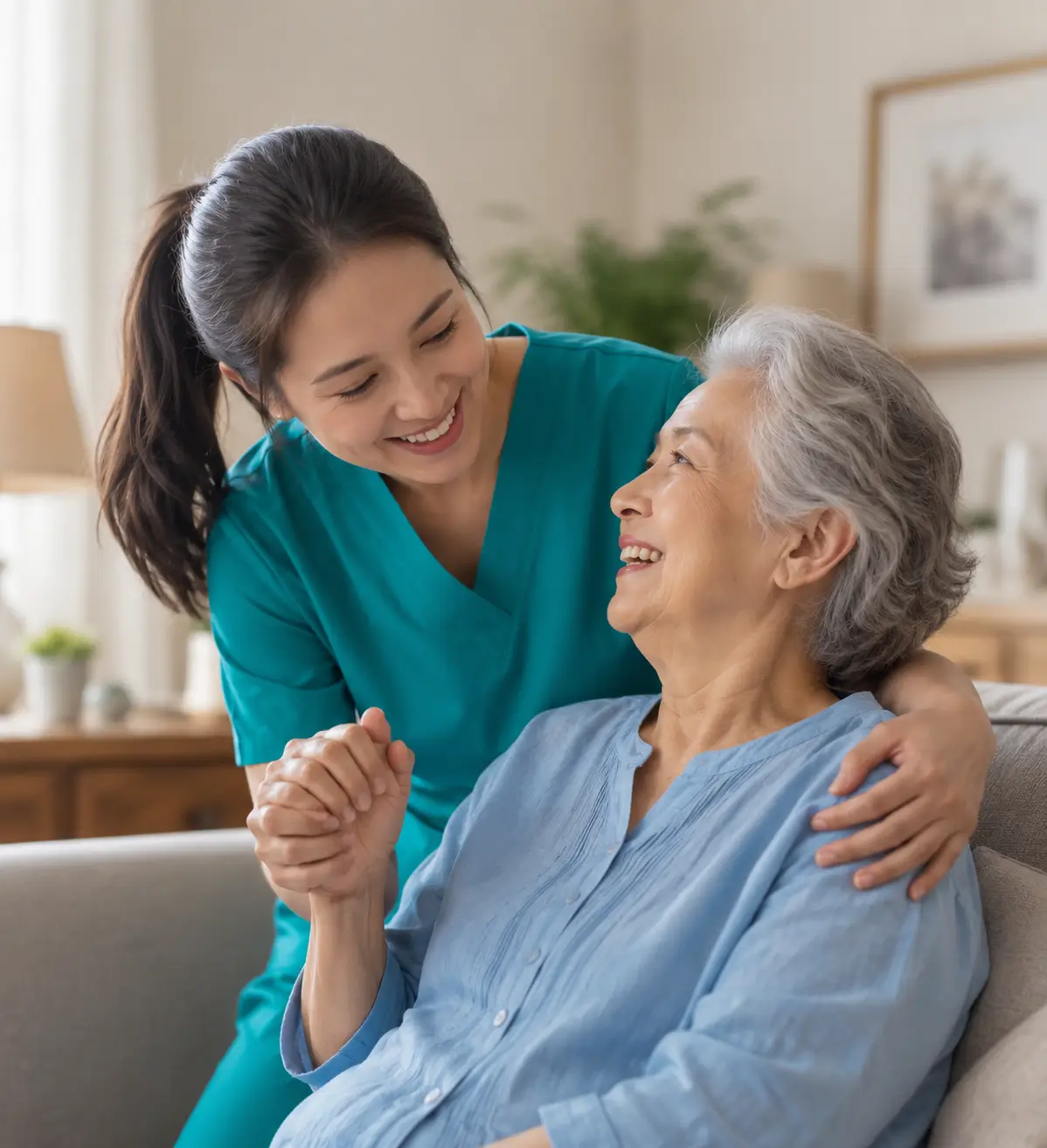 A smiling female caregiver in teal scrubs holding hands and supporting an elderly woman on a couch, representing compassionate dementia care in toronto.